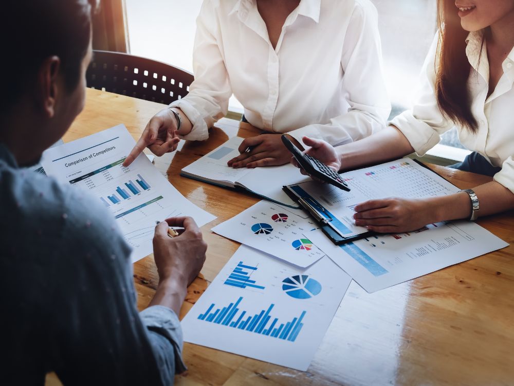 Reunião de equipe analisando relatórios financeiros e gráficos de desempenho sobre a mesa em ambiente corporativo.