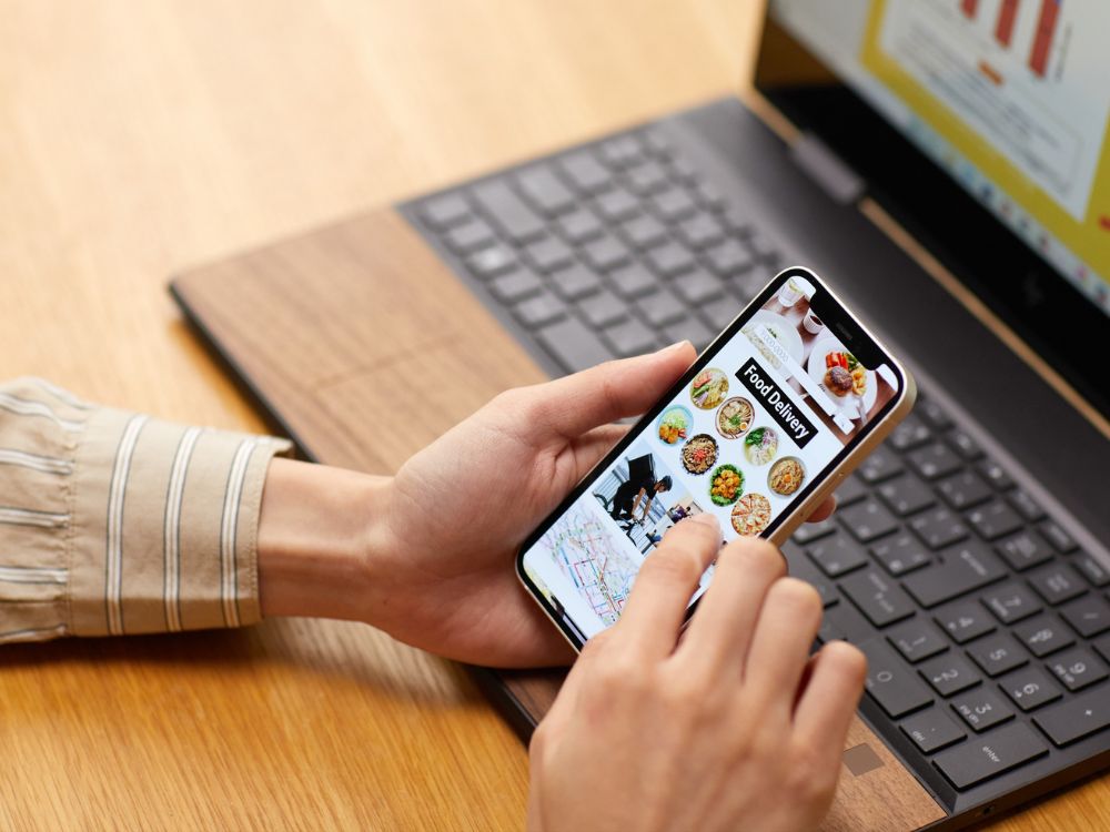 Pessoa utilizando smartphone com aplicativo de delivery aberto, enquanto trabalha no notebook sobre uma mesa de madeira, mostrando uso simultâneo de celular e computador.